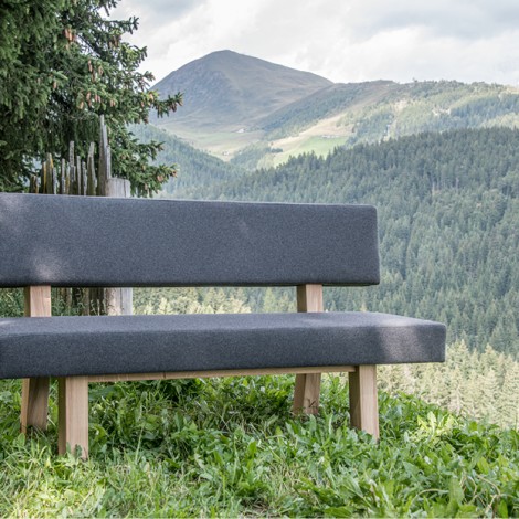 Gepolsterte Sitzbank mit Stützen aus Holz auf einer grünen Wiese vor dem Wald