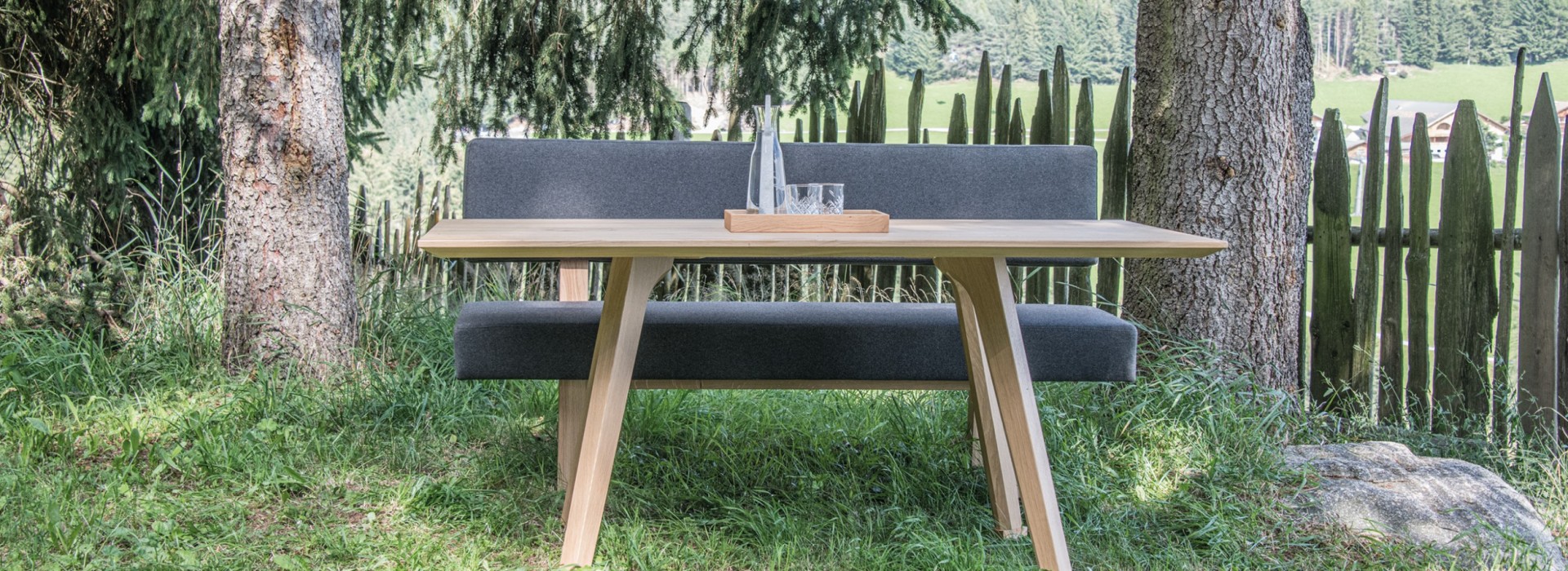 Sitzbank mit Tisch aus heimischem Holz auf einer grünen Wiese vor hohen Nadelbäumen