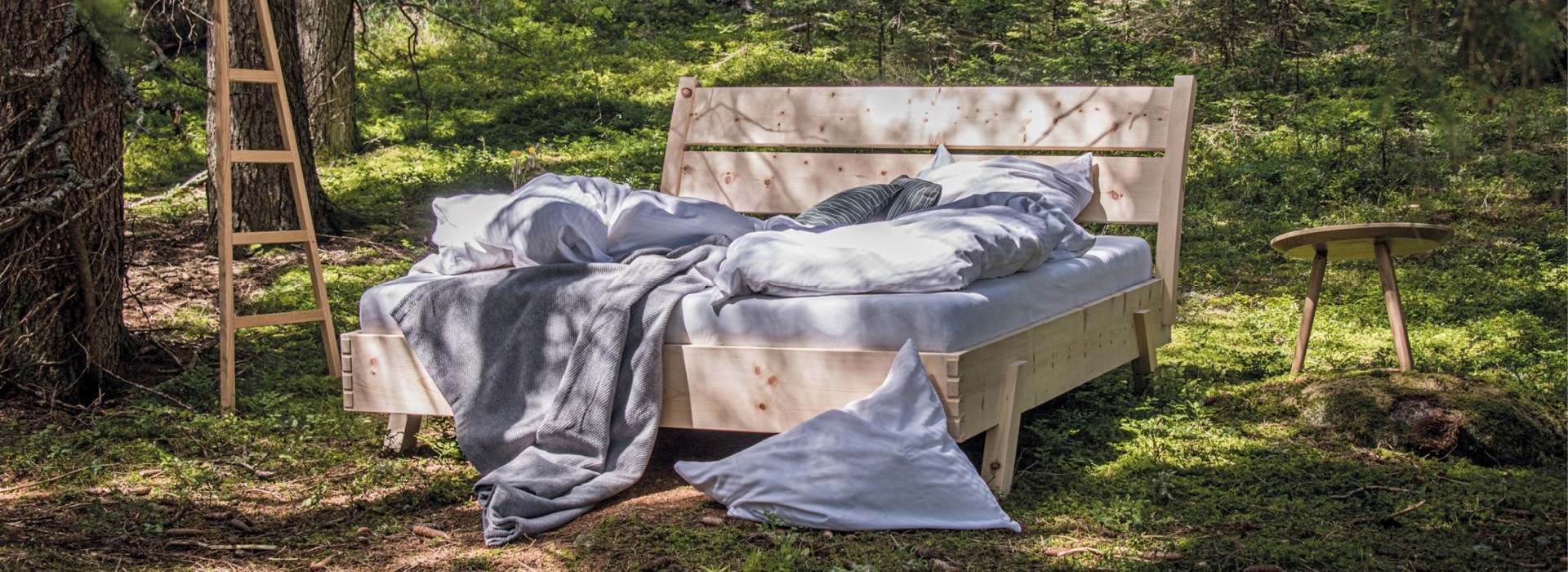 Stabiles Holzbett mit Rückenlehne, kleinem Holztisch und Leiterlampe inmitten der Natur im Wald