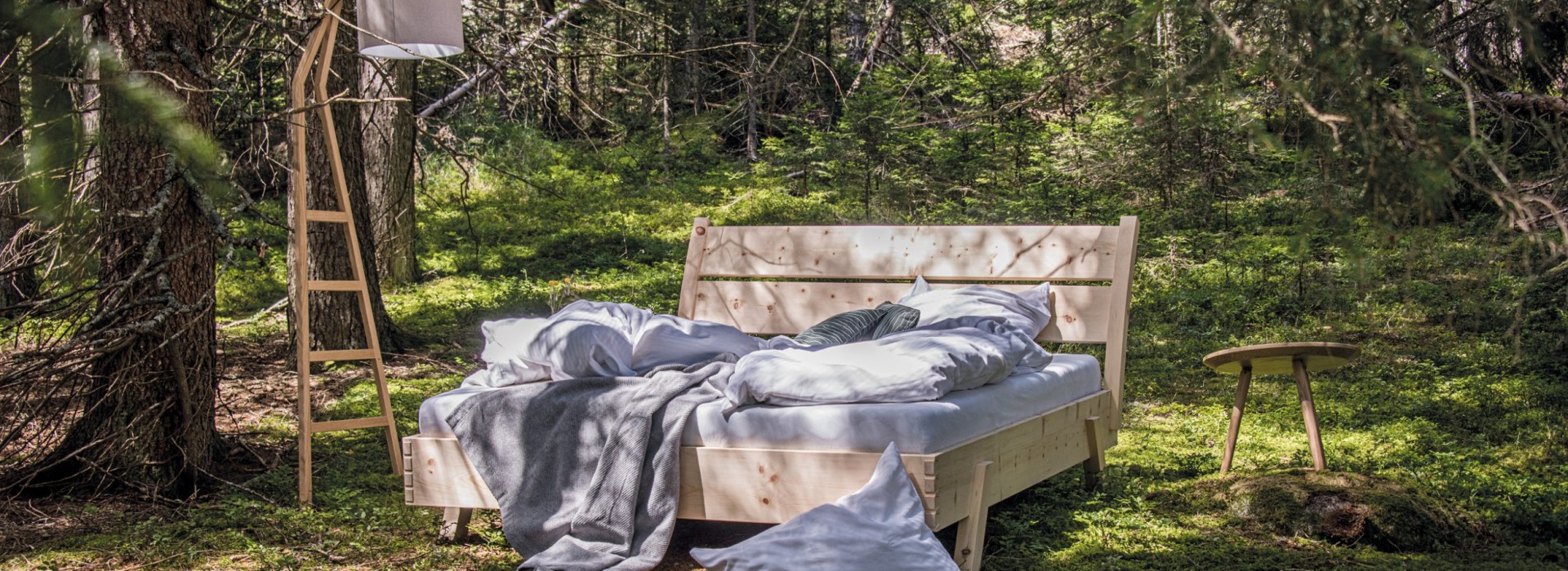 Holzbett mit Rückenlehne und Nachttisch mit Leiterlampe inmitten von Bäumen